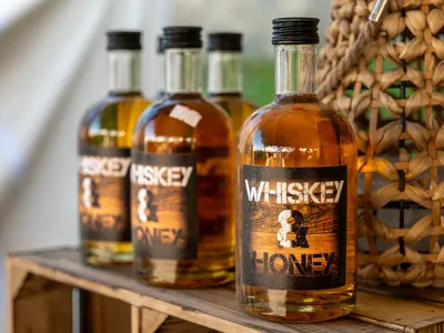 Vorschau: Mehrere Flaschen Whiskey & Honey auf einer Holzkiste neben einem geflochtenen Korb
